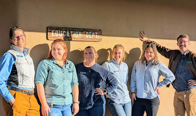 A group of six Day Camp Teachers standing in front of an adobe wall. The sun is shining and they are all smiling broadly.